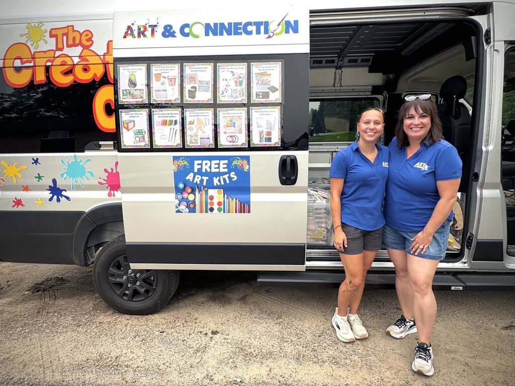 Two women in blue uniforms stand beside a white van with colorful artwork. The van has an open door displaying a sign that reads "FREE ART KITS."