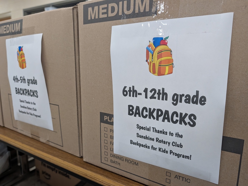 Boxes labeled "MEDIUM" and "6th-12th grade BACKPACKS" stacked on a shelf with signs attached to the front.