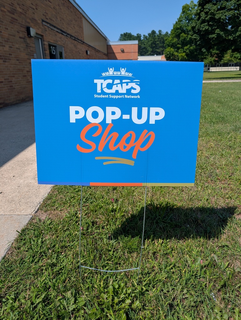 A blue sign on grassy lawn reads "TCAPS POP-UP SHOP." Behind it, a building and trees are visible.