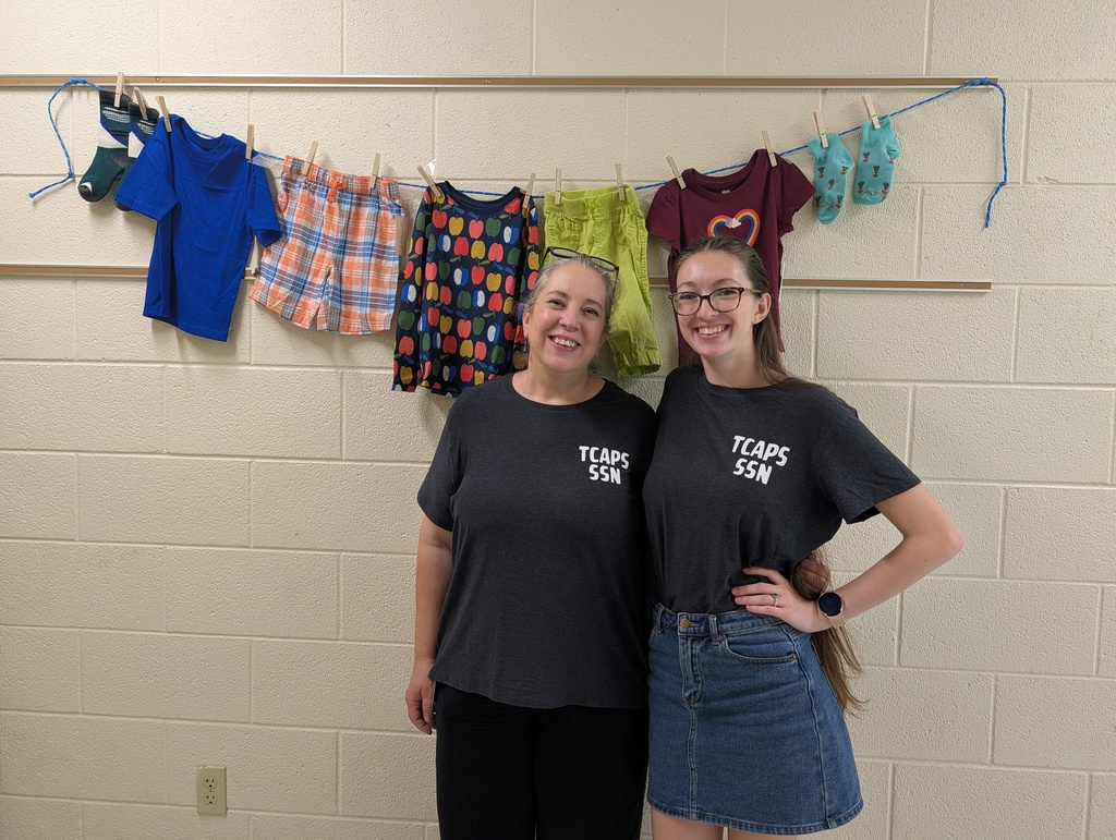 Two women stand in front of a white wall, wearing matching shirts, one with a denim skirt. Behind them, a line of clothes hangs on a rope.