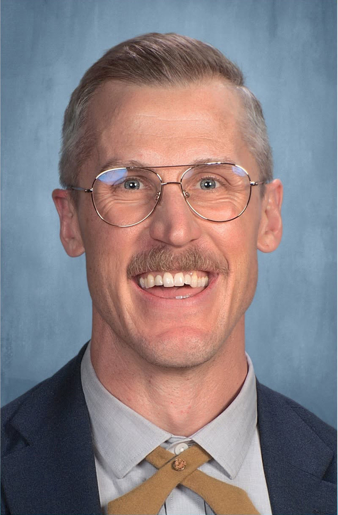 A man with short hair and a smile wearing glasses, a gray shirt, a brown tie, and a dark suit jacket.
