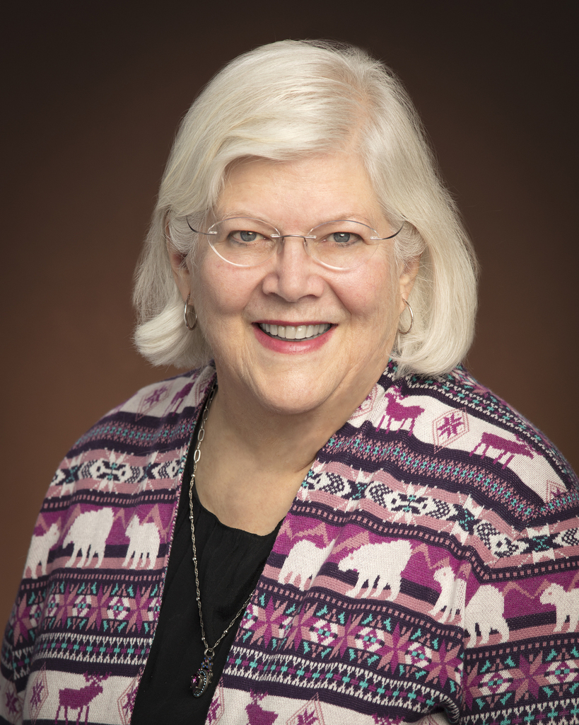 A smiling woman with white hair wearing glasses and a patterned jacket with polar bears stands against a dark background.