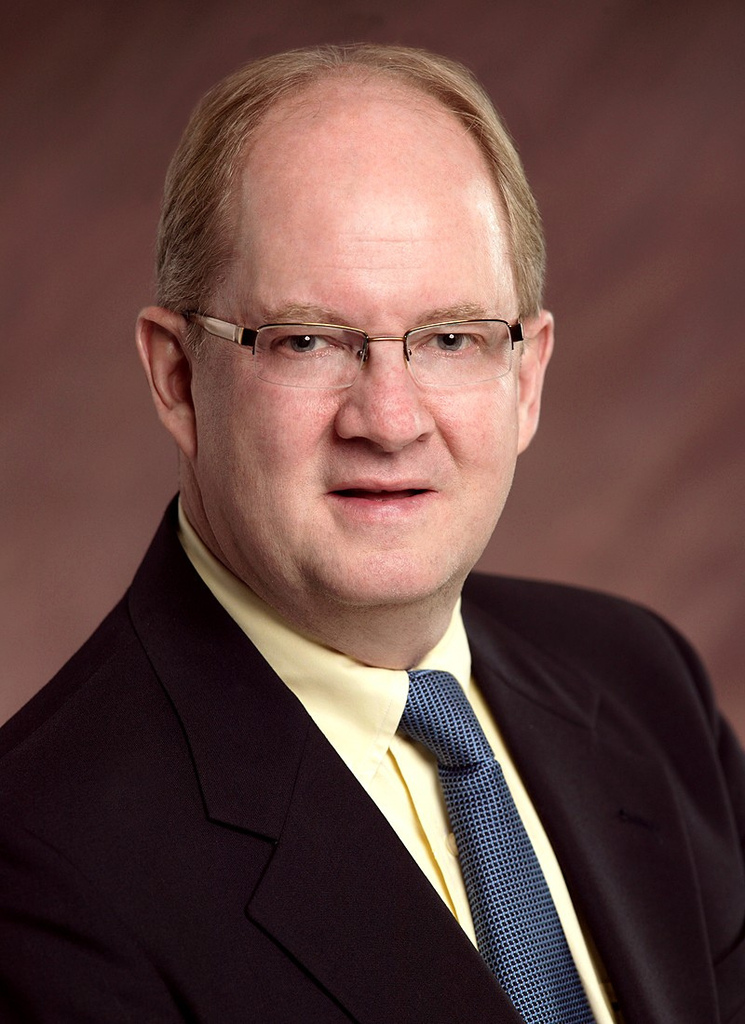 A man wearing a suit and tie with glasses on a brown background. He is looking at the camera.
