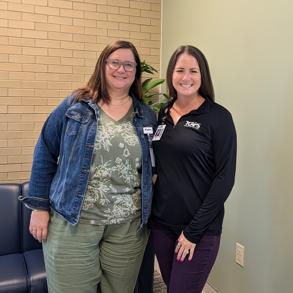 Two women standing, one in a denim jacket, another in a black shirt, in a room with a brick wall and a plant.