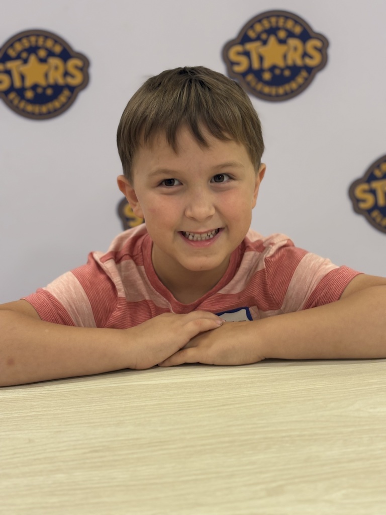 A young boy in a pink and white striped shirt smiles at the camera, seated at a table. Behind him is a wall with logos.