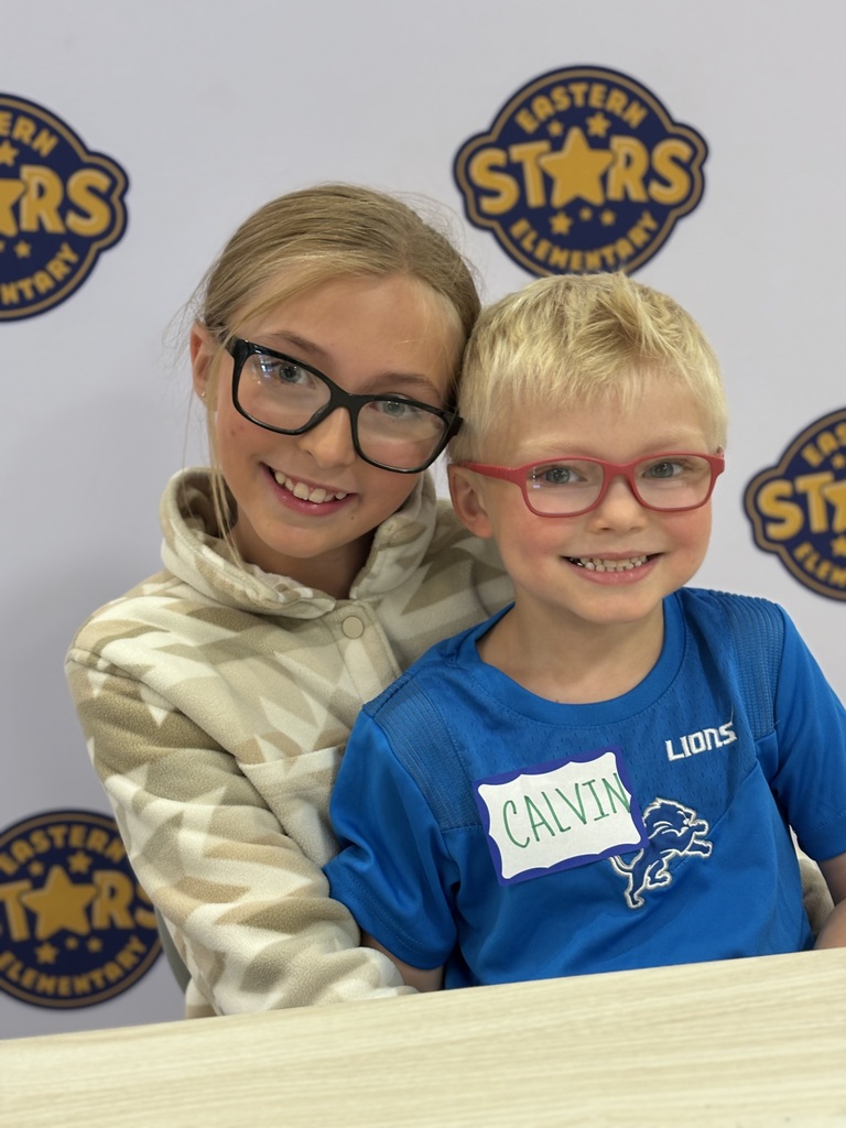 Two kids, one girl and one boy, sit at a desk wearing glasses. The boy wears a blue shirt.