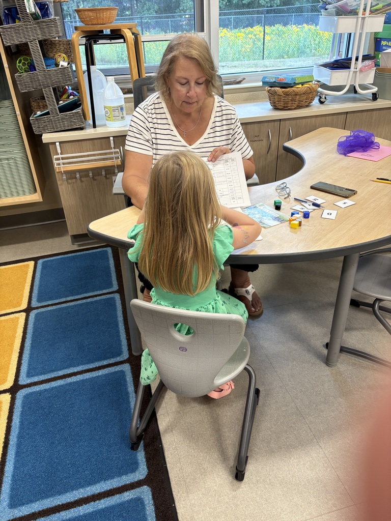 Woman and child at a desk. Child is seated, woman is standing behind her. They both appear to be reading.