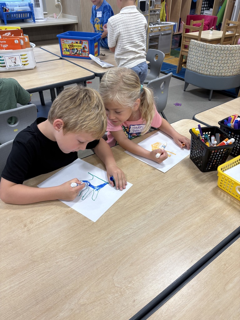 Two children sitting at a table drawing with markers. Background includes other desks with people.