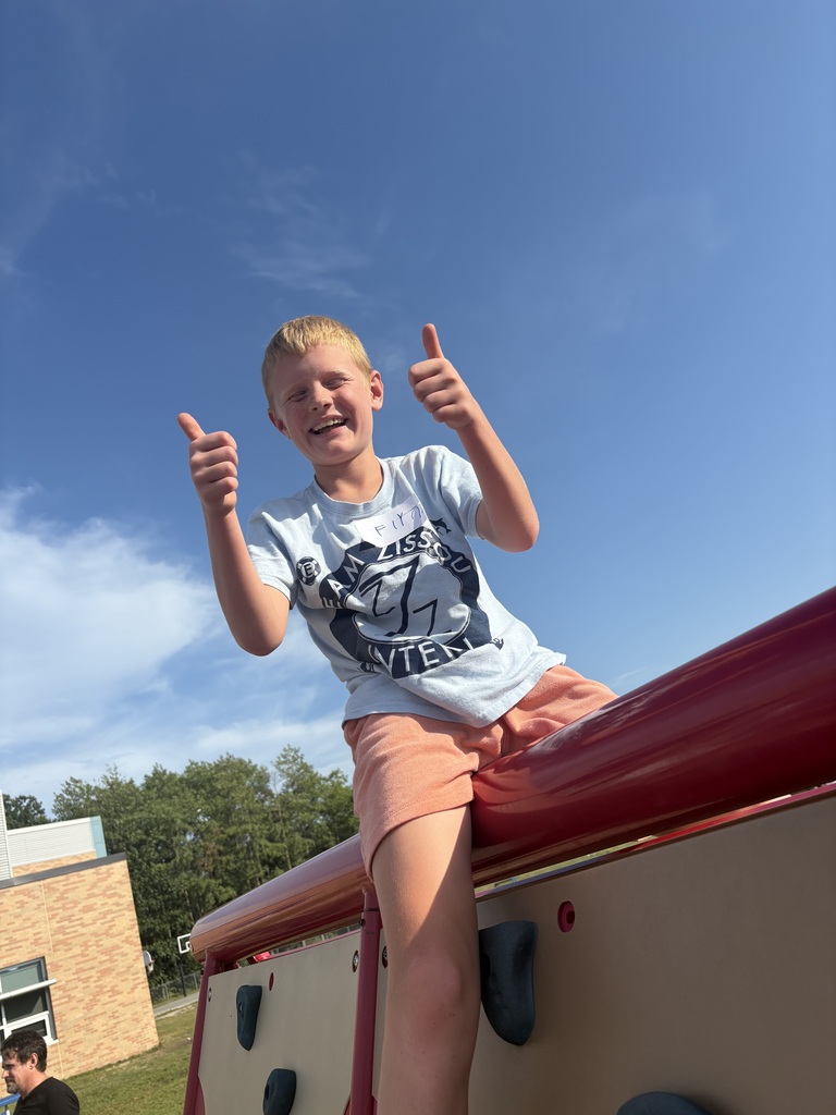 A child in a white T-shirt and orange shorts smiles and gives a thumbs-up, sitting on a playground structure.