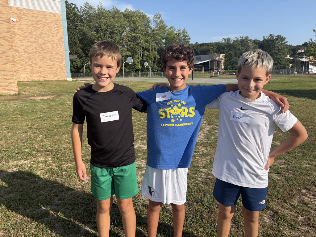 Three boys stand together on a grassy field. They wear different colored shirts and shorts. Behind them is a building and trees.