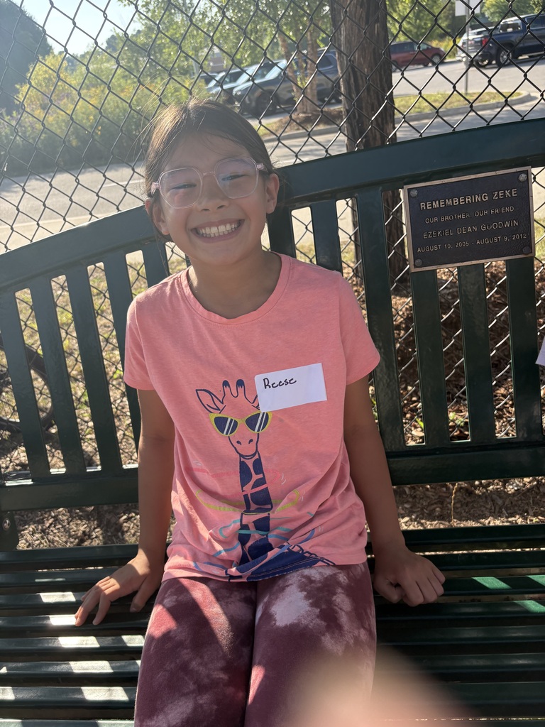A child in a pink shirt with a giraffe print and glasses smiles at the camera. Behind them is a green bench with a plaque.