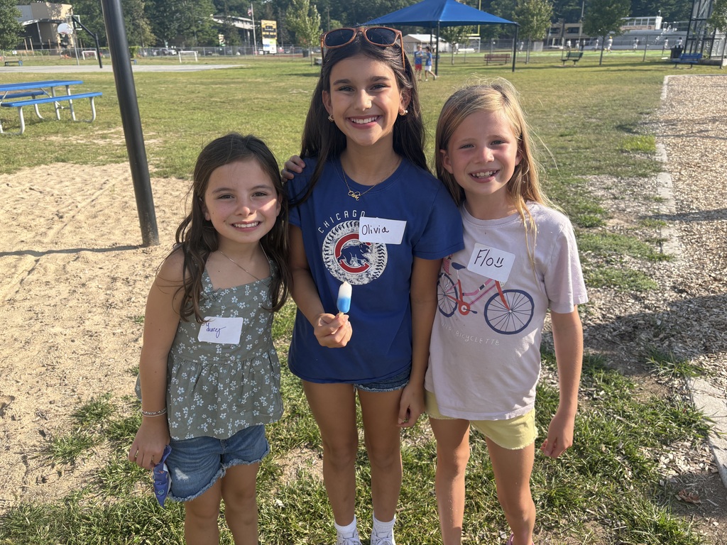 Three girls in outdoor area, holding a popsicle. One wears a name tag. A bench is behind them.