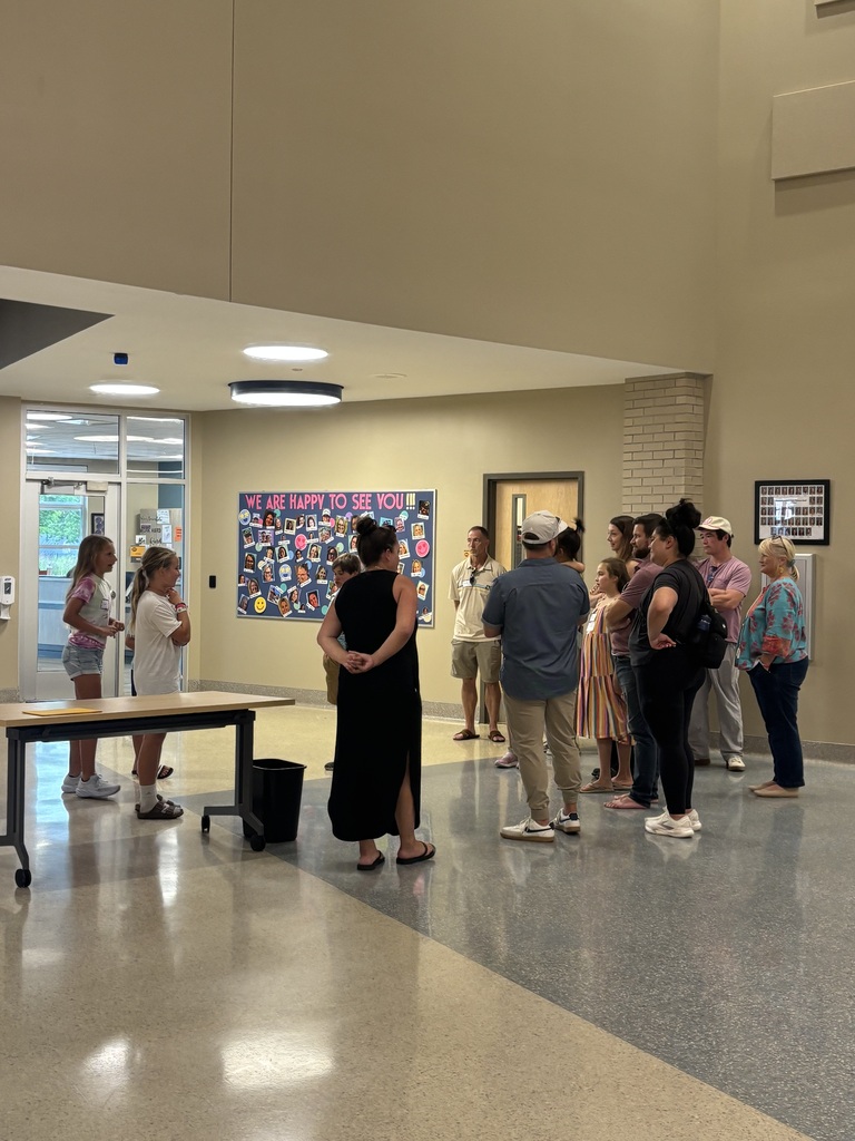 Several people stand in a hallway around a table. The walls have artwork, and overhead lights are on.