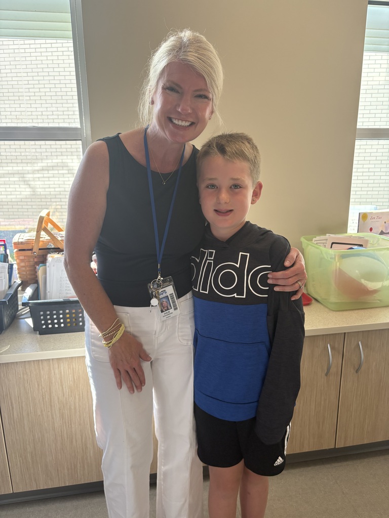 Woman and boy in school environment, smiling at camera, standing in front of wooden cabinets and window.
