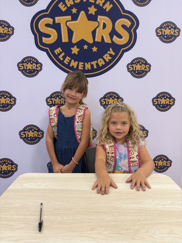 Two young girls sit at a table with pens in front of a wall with the words "Eastern Stars Elementary".