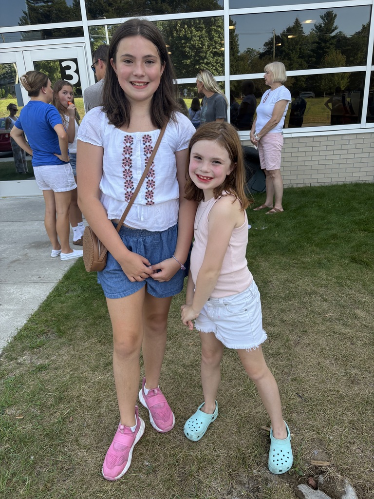 Two young girls in casual clothes stand in front of a building with glass walls, with more people in the background.