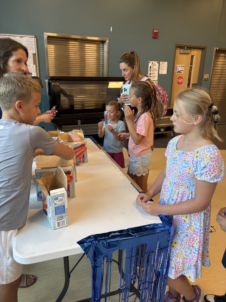 Children gather around a table with food items in a room. A boy packs a box, and a girl watches.