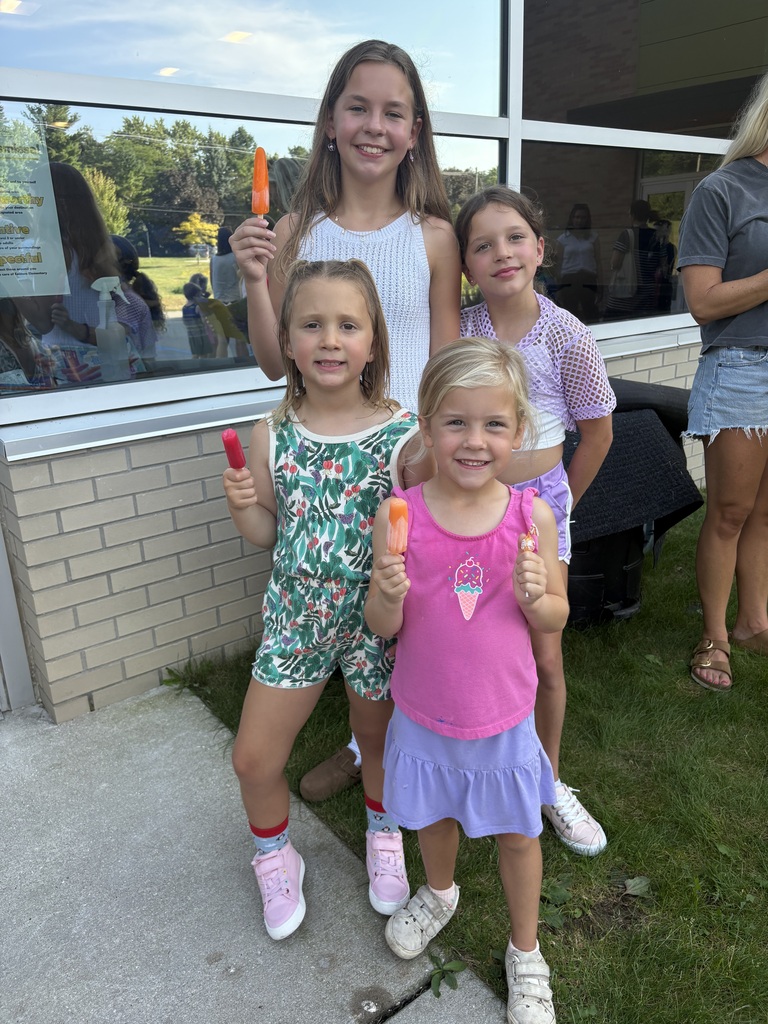 Four young girls holding ice pops stand on a sidewalk outside a building. One wears a white dress.