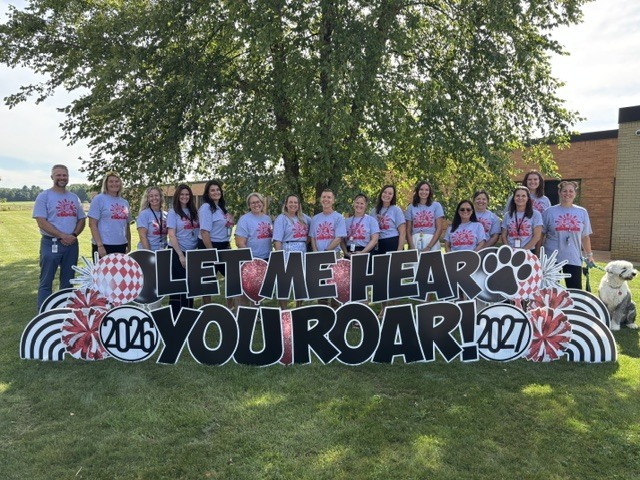 A group wearing white T-shirts poses near a sign saying "LET ME HEAR YOU ROAR!" on a grassy field with trees.