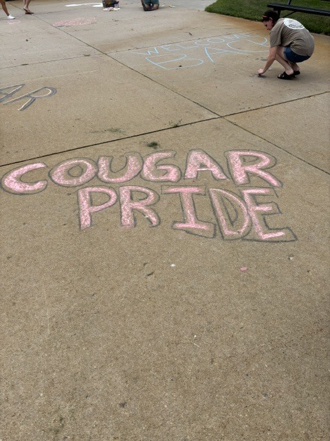 Person kneels, chalking "COUGAR PRIDE" on concrete, others in background. Sidewalk, grassy area.