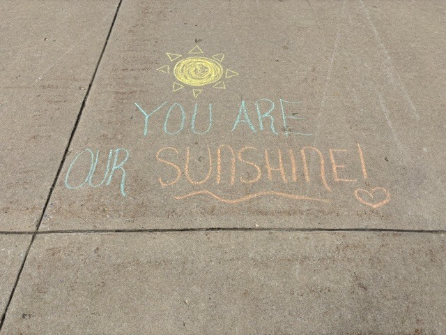Sidewalk with chalk drawing: a sun with the words "You are our sunshine!" in blue and orange.