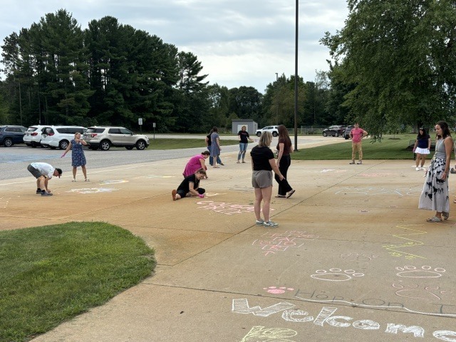 People in various clothing stand and crouch on a concrete pad, creating chalk drawings that read "Welcome." Trees and parked cars are visible in the background.