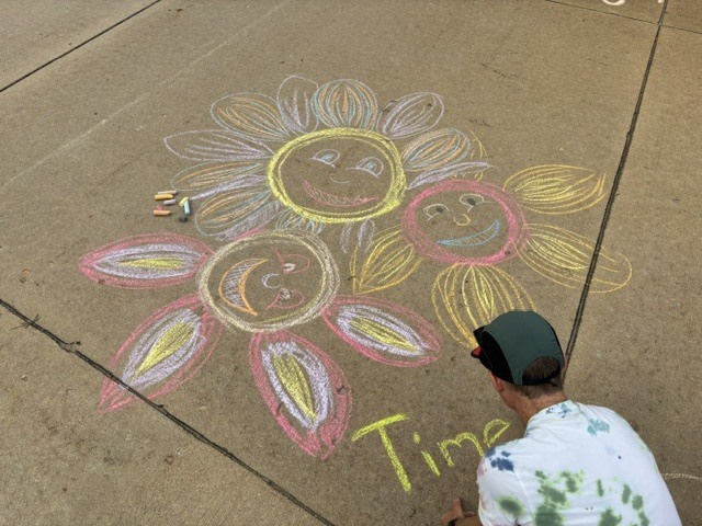 A person kneels, drawing a flower and three smiling faces on a sidewalk. The word "Time" is written nearby.
