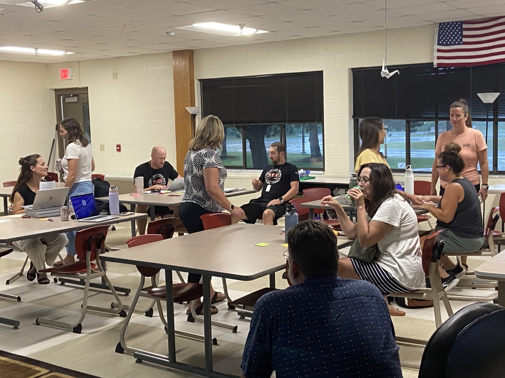 Several people are gathered in a meeting room. Some sit at tables with laptops, while others stand and talk. An American flag hangs on the wall.