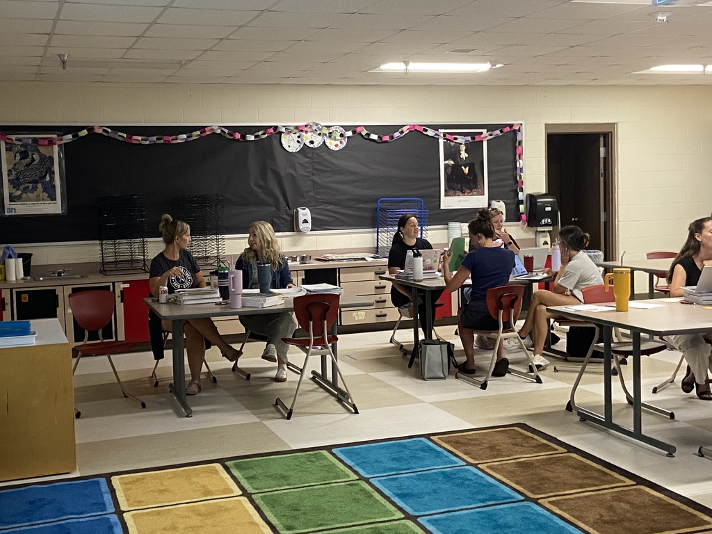 A group of people seated at tables in a classroom with a bulletin board, a rug, and desks.