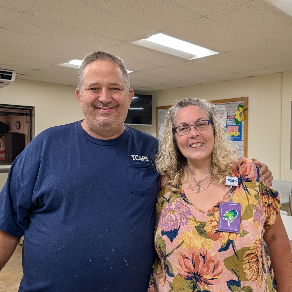 A man and woman standing close, smiling, in a room with white walls and tiled flooring.