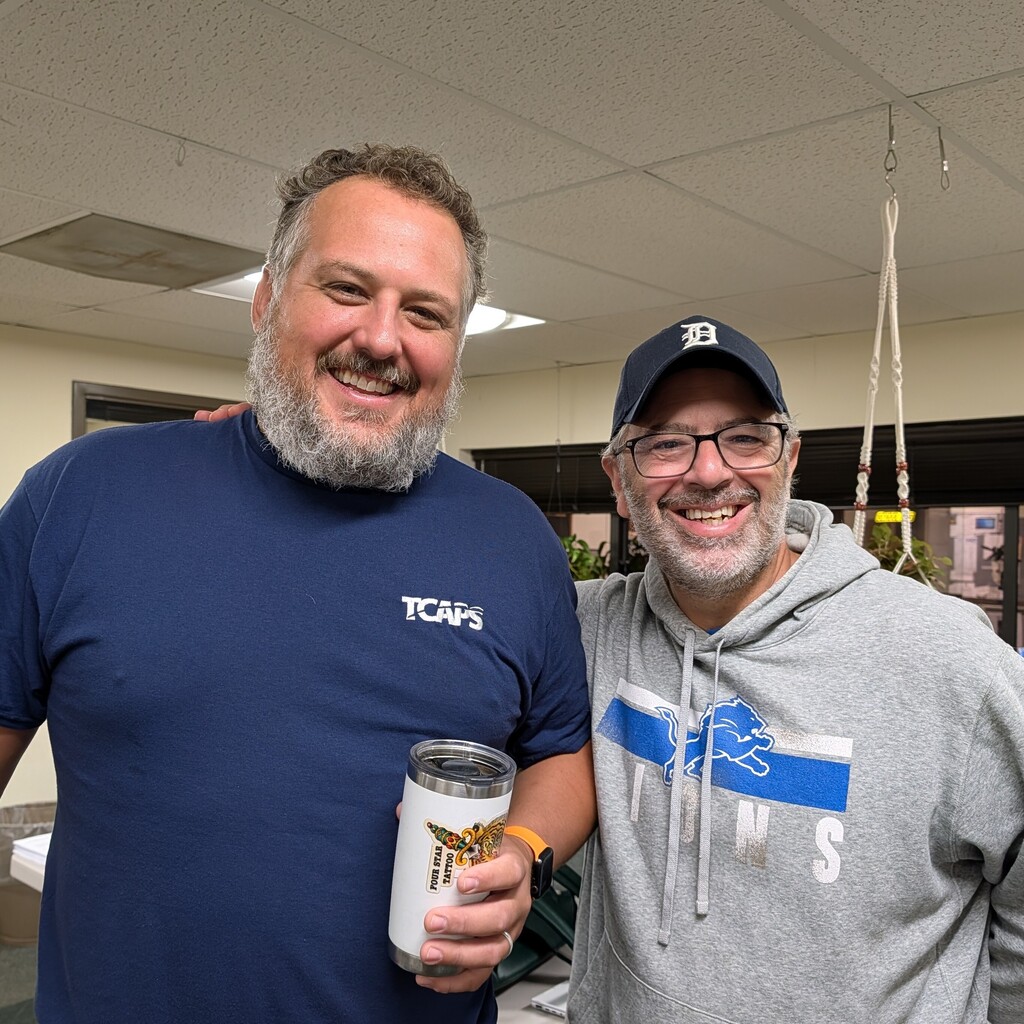 Two men, one in a blue shirt and one in a gray hoodie, standing together and smiling.
