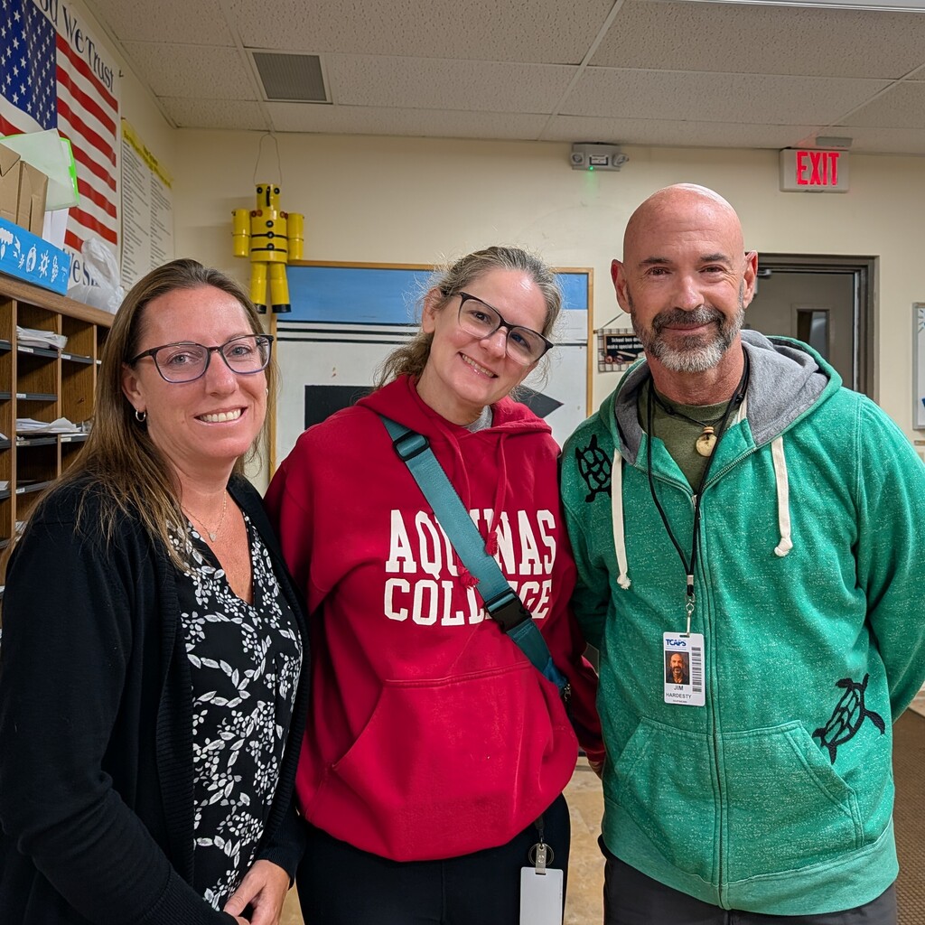 Three adults pose for a photo in a room with shelves, a flag, and an exit sign.