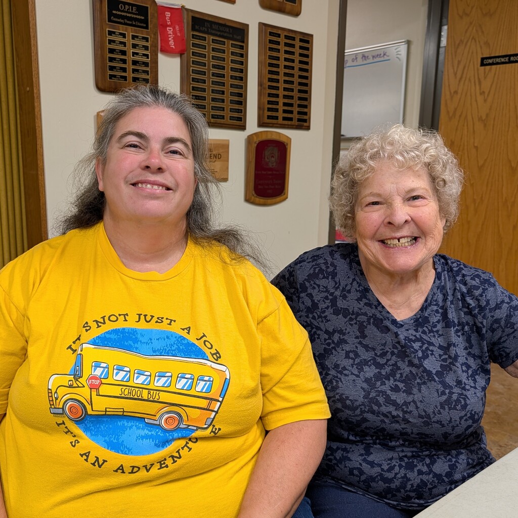Two women smiling and posing for a photo; one wears a yellow shirt with a school bus design.