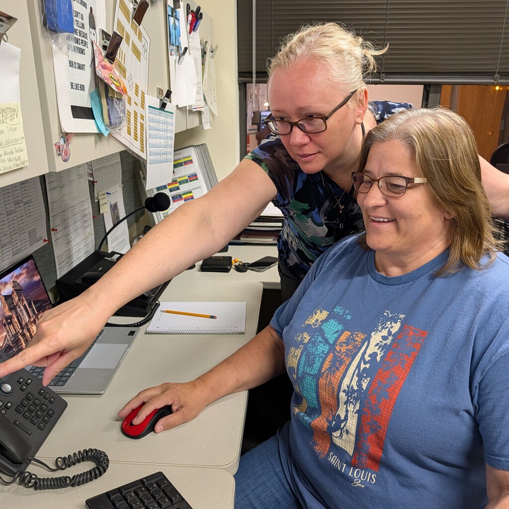 Two women in an office setting; one seated with a blue t-shirt, the other standing behind her.