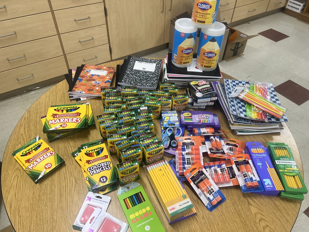 A wooden table filled with various school supplies, including notebooks, pencils, markers, and crayons, against a backdrop of wooden cabinets.