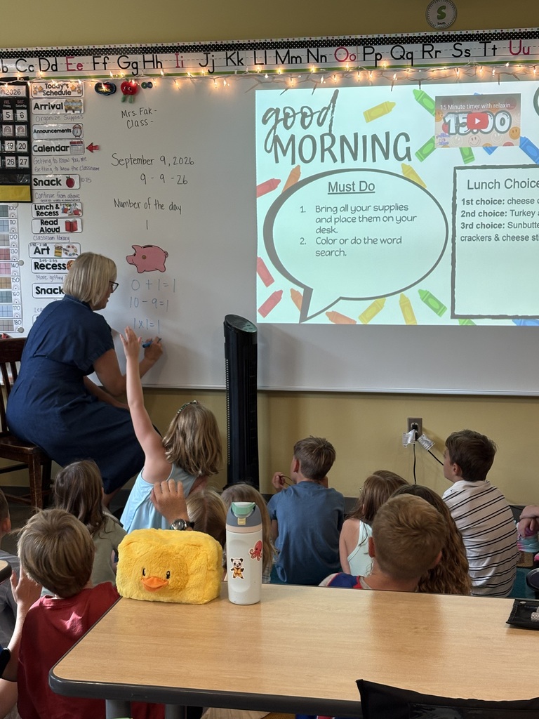 A classroom with a teacher writing on a whiteboard. Children are seated on the rug in front of a table with a yellow toy.