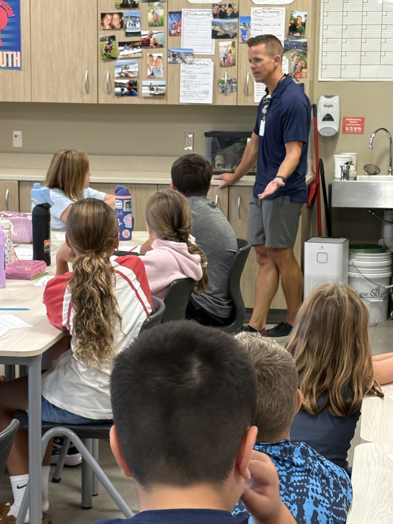 A classroom with students seated around tables. A teacher stands at the front of the room, facing the students.