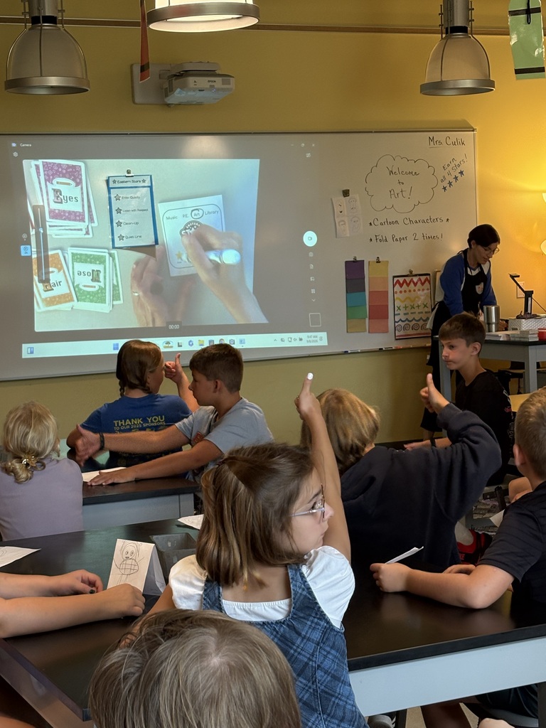 Students in a classroom attentively watching a presentation projected on a screen, seated at tables.