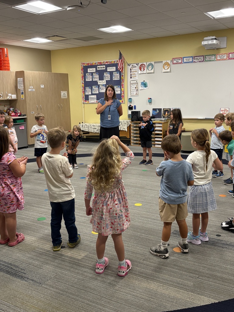 In a classroom, children form a circle around a woman who is standing. The room has a whiteboard, flags, and chairs.