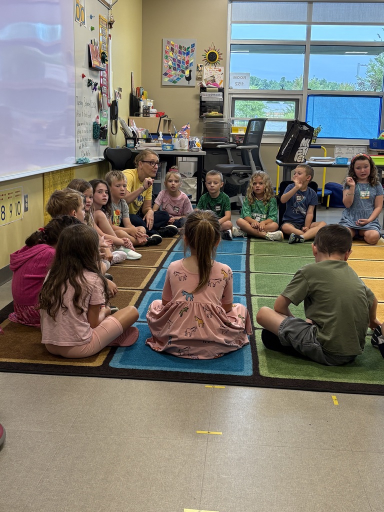 Children sit on rugs in a classroom, facing a teacher. A whiteboard and bulletin board are visible.