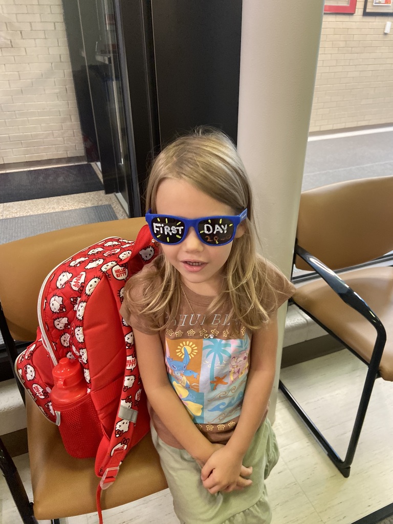 A child with a red backpack sits in a chair, wearing blue sunglasses and a colorful shirt. Behind, two chairs and a glass door are visible.