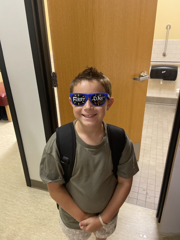 A young boy stands in a hallway with a smile. He wears a backpack and blue glasses. A wooden door and tiled floor are behind him.
