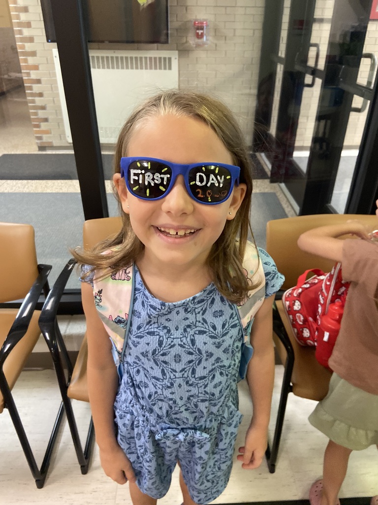 A young girl with blonde hair, wearing blue overalls and sunglasses that read "First Day." She smiles while standing in a room with chairs and a door.