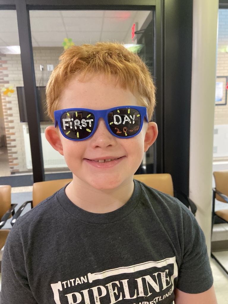 A child with red hair smiles while wearing blue sunglasses with the words "First Day" on them.
