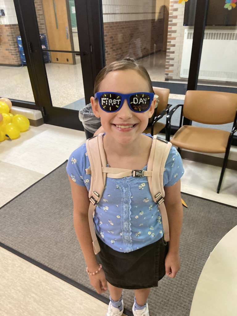 A young girl wearing sunglasses with "First Day" text stands in a room with chairs and a rug.