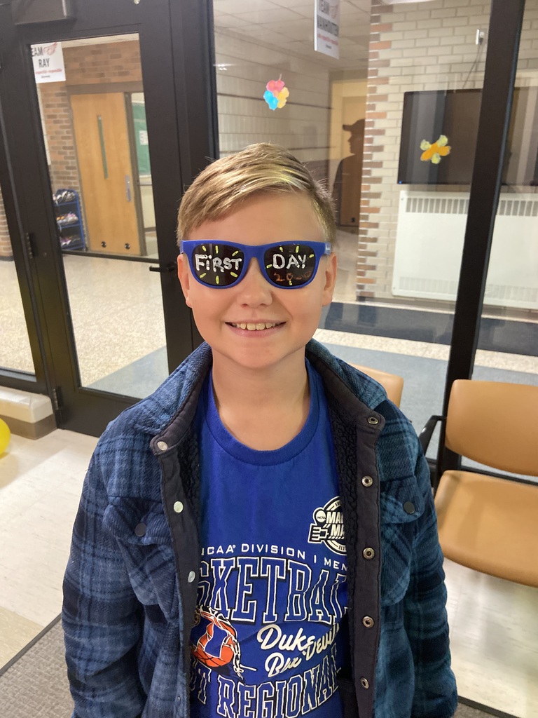 A young boy wearing a blue jacket and a blue shirt stands indoors, smiling with a pair of glasses on his face.