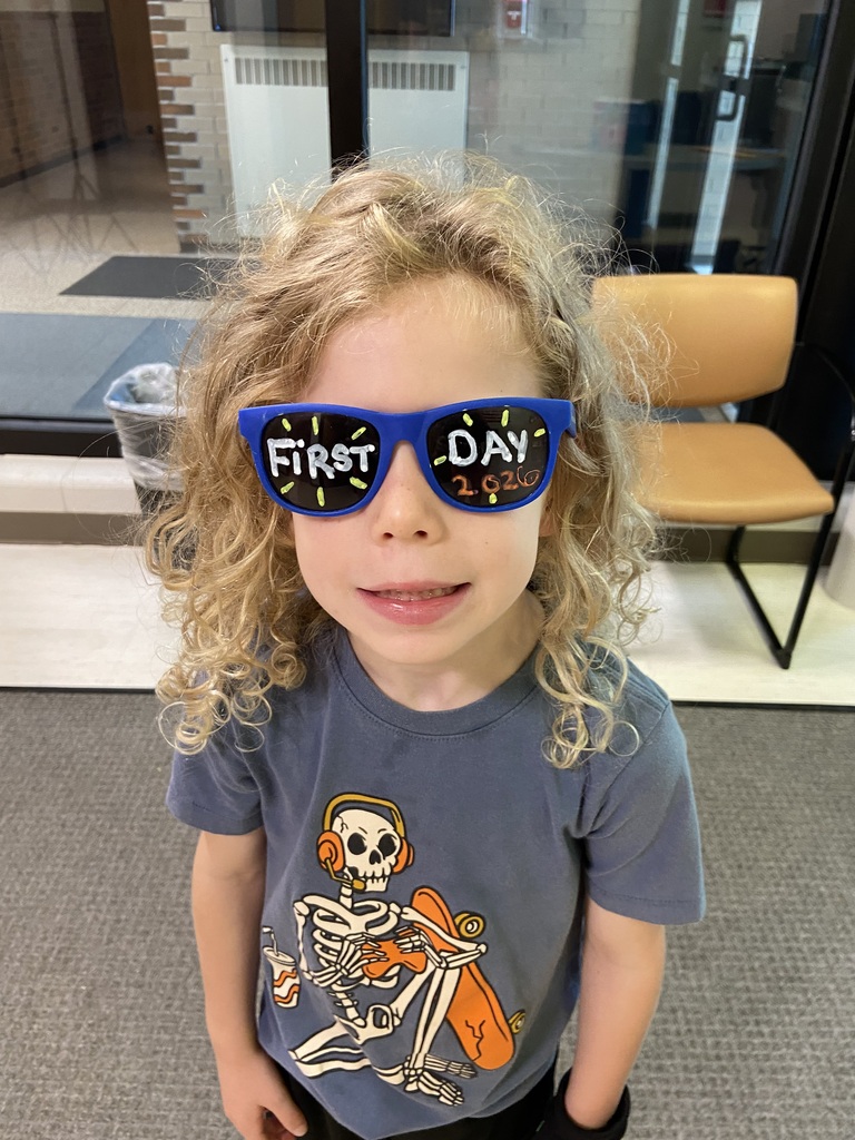 A child with blonde hair stands in a room with blue glasses, wearing a gray T-shirt with a skeleton design.