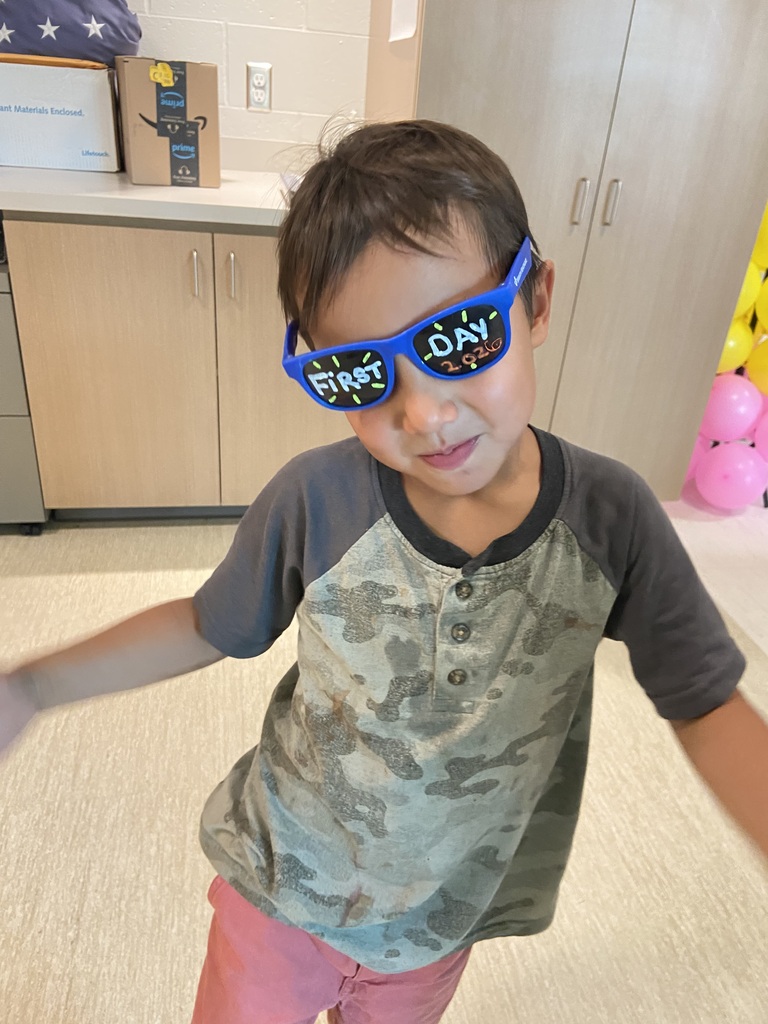 A child in a camouflage shirt, pink pants, and blue "First Day" sunglasses is standing. Behind is a kitchen area with cabinets and balloons.