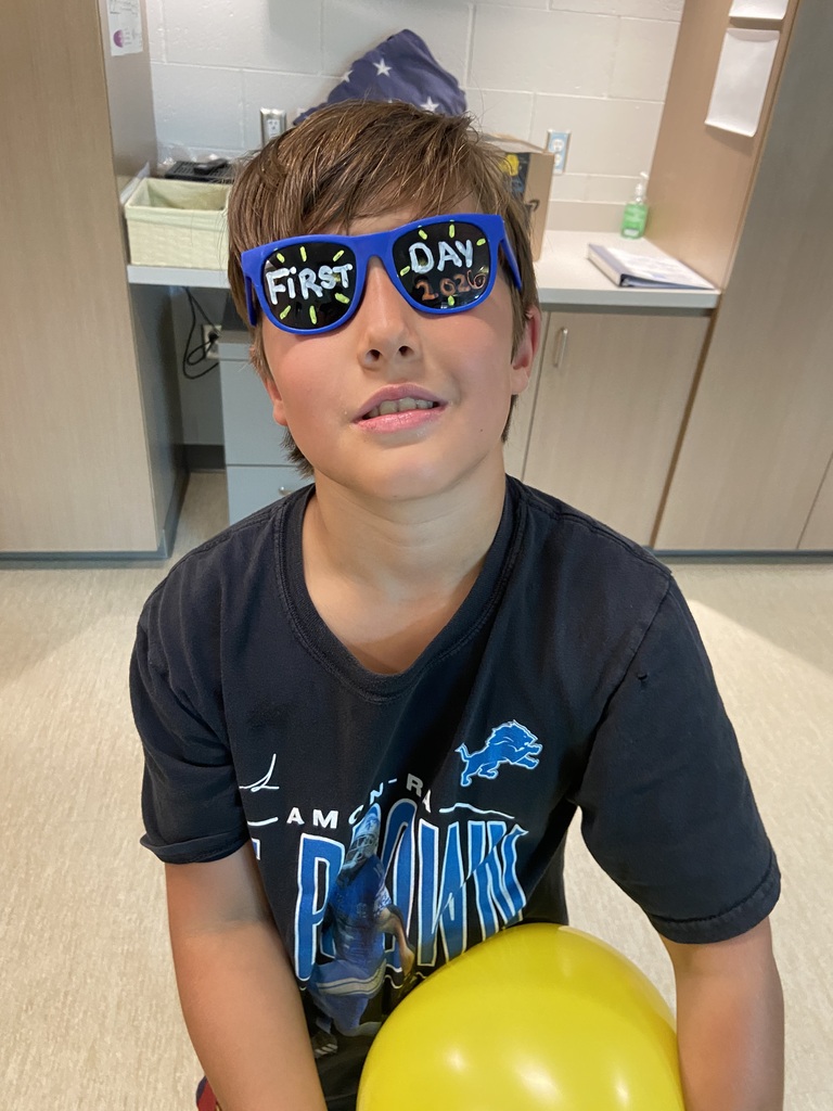A young boy wearing a blue shirt with a graphic, holding a yellow balloon, and wearing blue sunglasses with white letters.