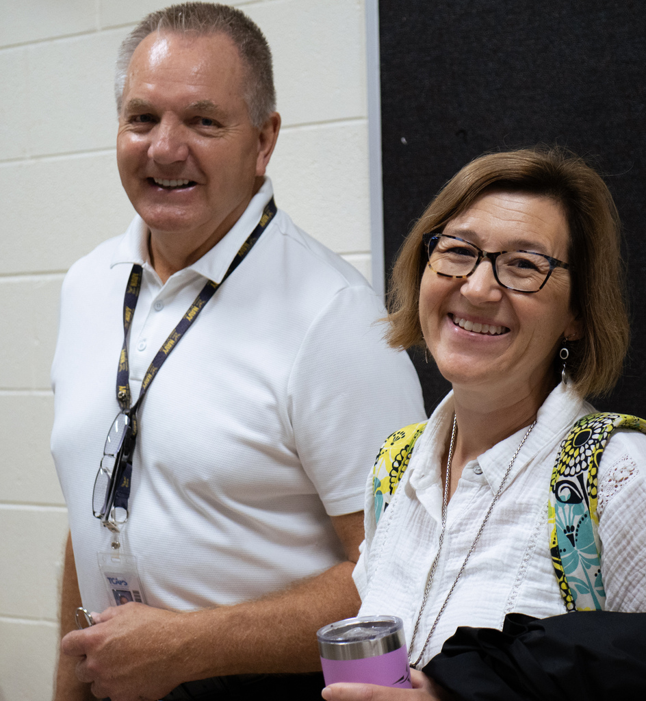 Two people stand in a room, smiling. A man wears a white shirt and glasses on a lanyard. A woman holds a purple cup.